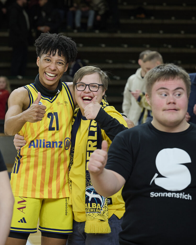 Deeshyra Thomas, die Profi-Spielerin von Alba Berlin, nimmt den Sonnenstich-Artisten Milan Godding in den Arm. Im Vordergrund freut sich Sonnenstich-Artist Luzian Bücker.