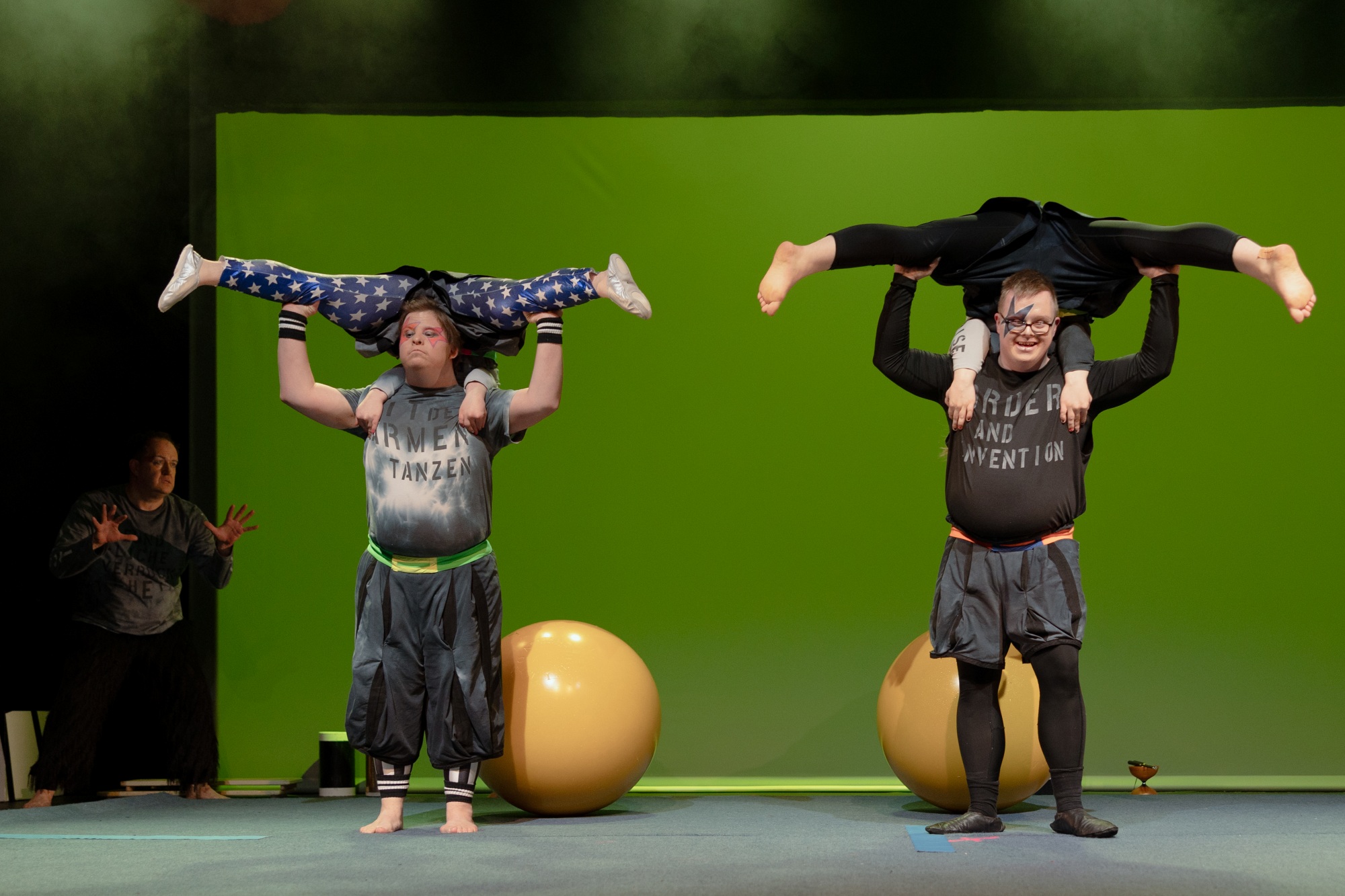 Vor grünem Hintergrund und vor zwei gelben Kugeln tragen die Artisten Sascha Perthel und Oskar Schenck die Artistinnen Sarah Walther und Olivia Wolff auf den Schultern