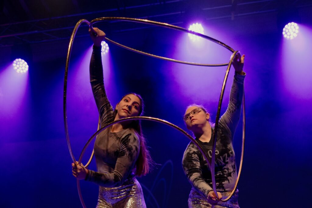 Die Artistinnen Asil Ashour und Anne-Sophie Mosch sind vor blauem Hintergrund bei ihrer Hoop-Nummer in Nahaufnahme mit vier Hoops zu sehen.