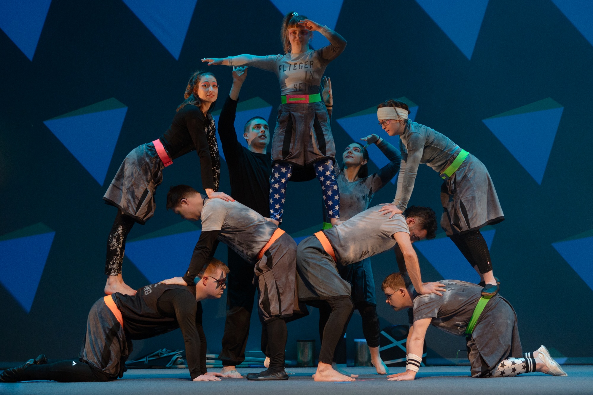 Hier zeigen 9 Artistinnen und Artisten Circus Sonnenstich eine Bodenakrobatik in Form einer Pyramide. Ganz oben auf den Schultern ist Sarah Walther zu sehen. Auf ihrem Kostüm steht "Fliegerin sein".