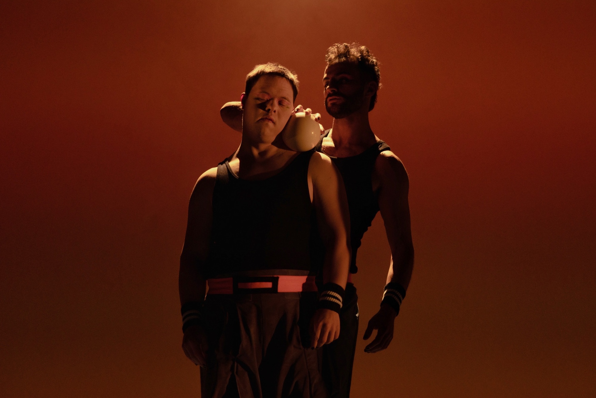 Vor braun-rotem Hintergrund, der an einen Sonnenuntergang erinnert, sind Jannic Golm und Alaa Abu Alrob in schwarzen Kostümen mit einer weißen, handgroßen Kugel zu sehen. Sie stehen nahe zusammen. Das Foto hat eine sehr intime Atmosphäre