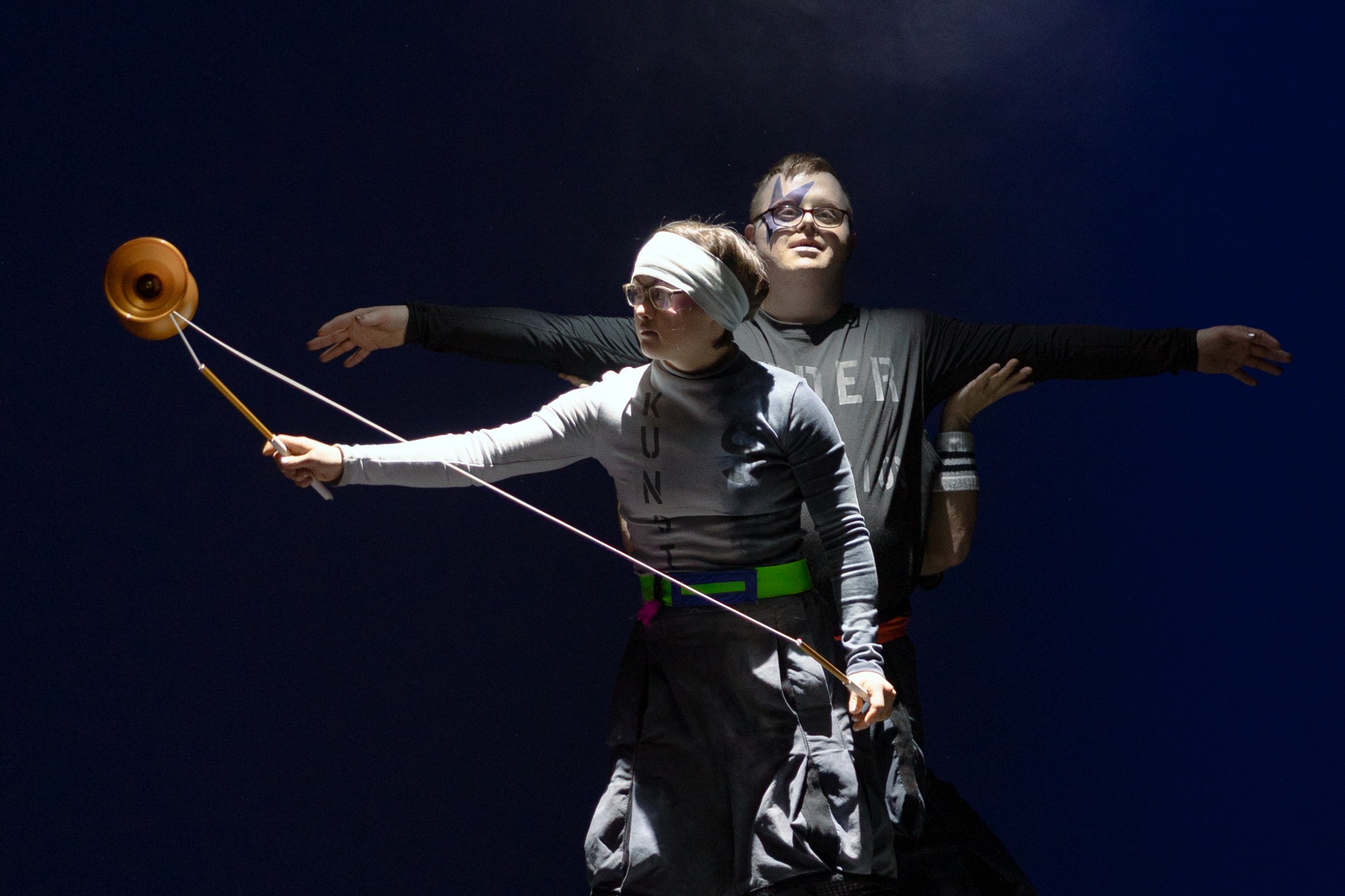 Sol-Rein Saunders und Oskar Schenck zeigen vor dunkelblauem Hintergrund eine gemeinsame Diabolo-Nummer. Ihre Gesichter sind in helles Licht getaucht.