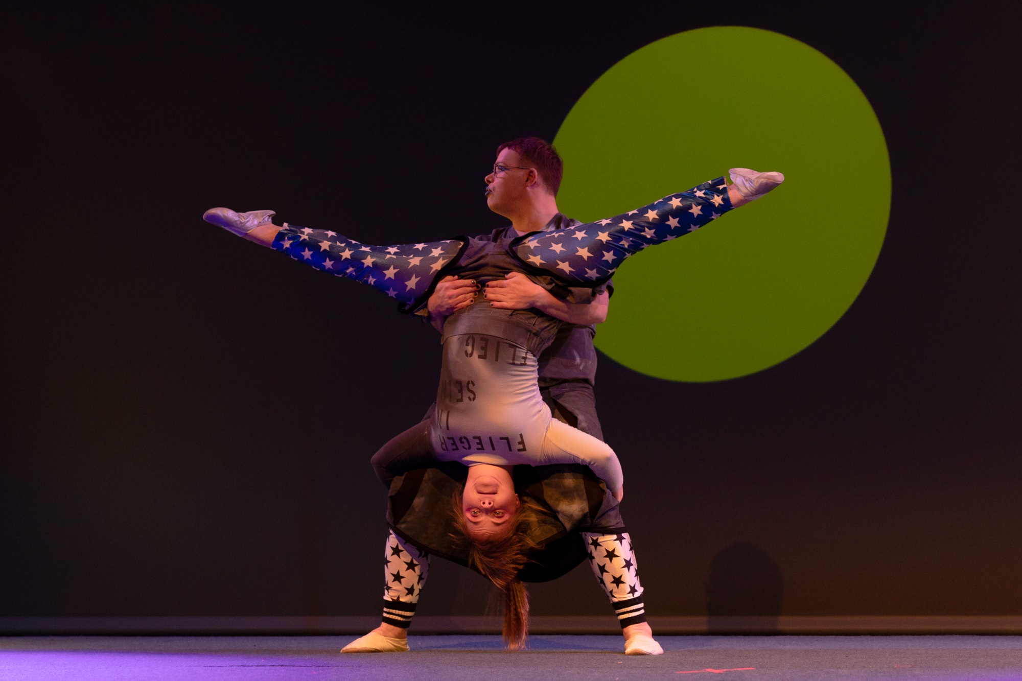 Auf dem Foto zu sehen sind die Artisten Florian Klotz und Sarah Walther bei ihrer Partnerakrobatik. Florian hält Sarah an der Hüfte. Sarah hängt dabei kopfüber, schaut nach vorne und hält sich an Florians Kniekehlen fest.