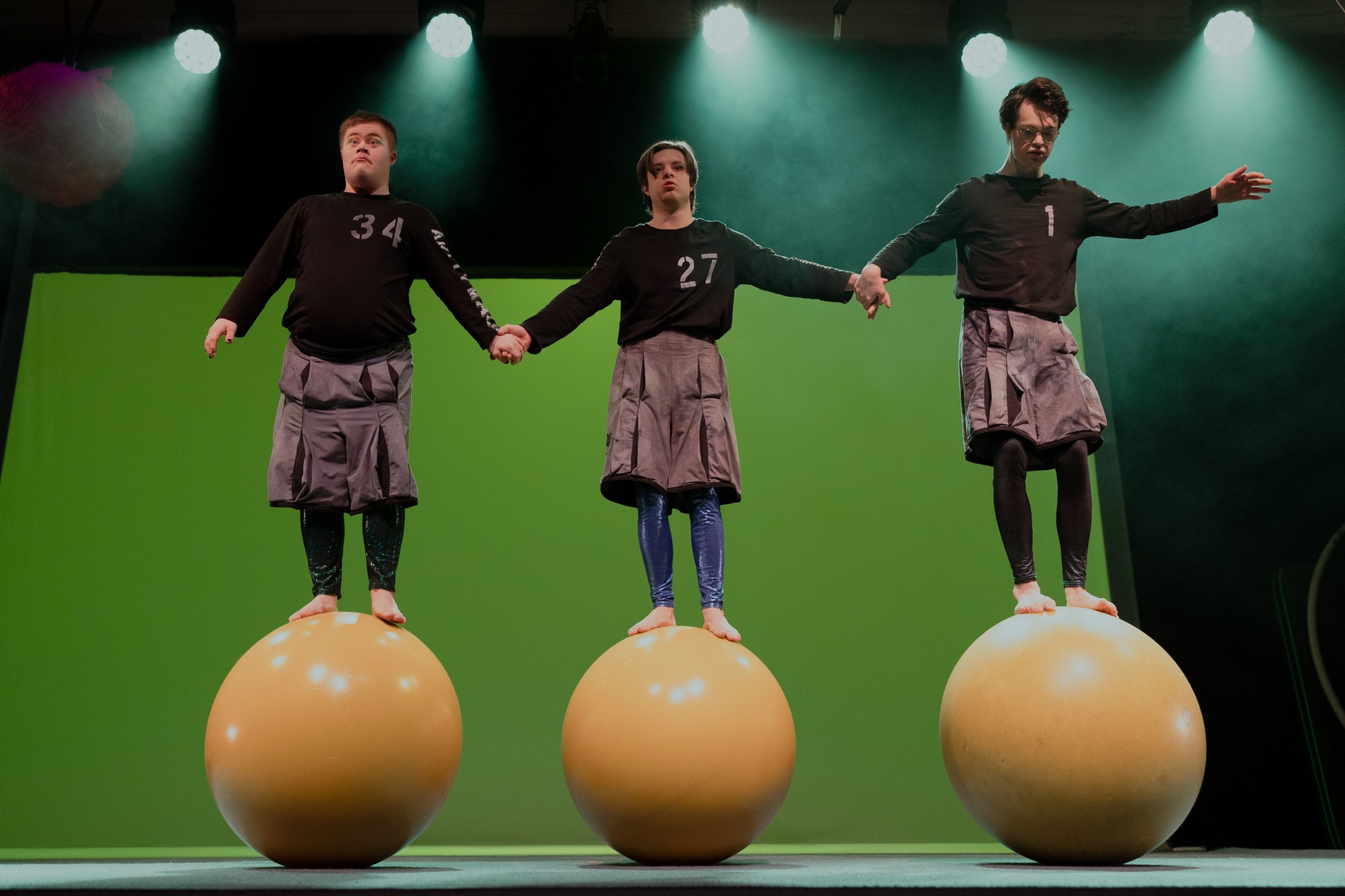 Auf gelben Laufkugeln vor froschgrünem Hintergrund stehen die Artisten Luzian Bücker, Neon Spohn und Elias Massing